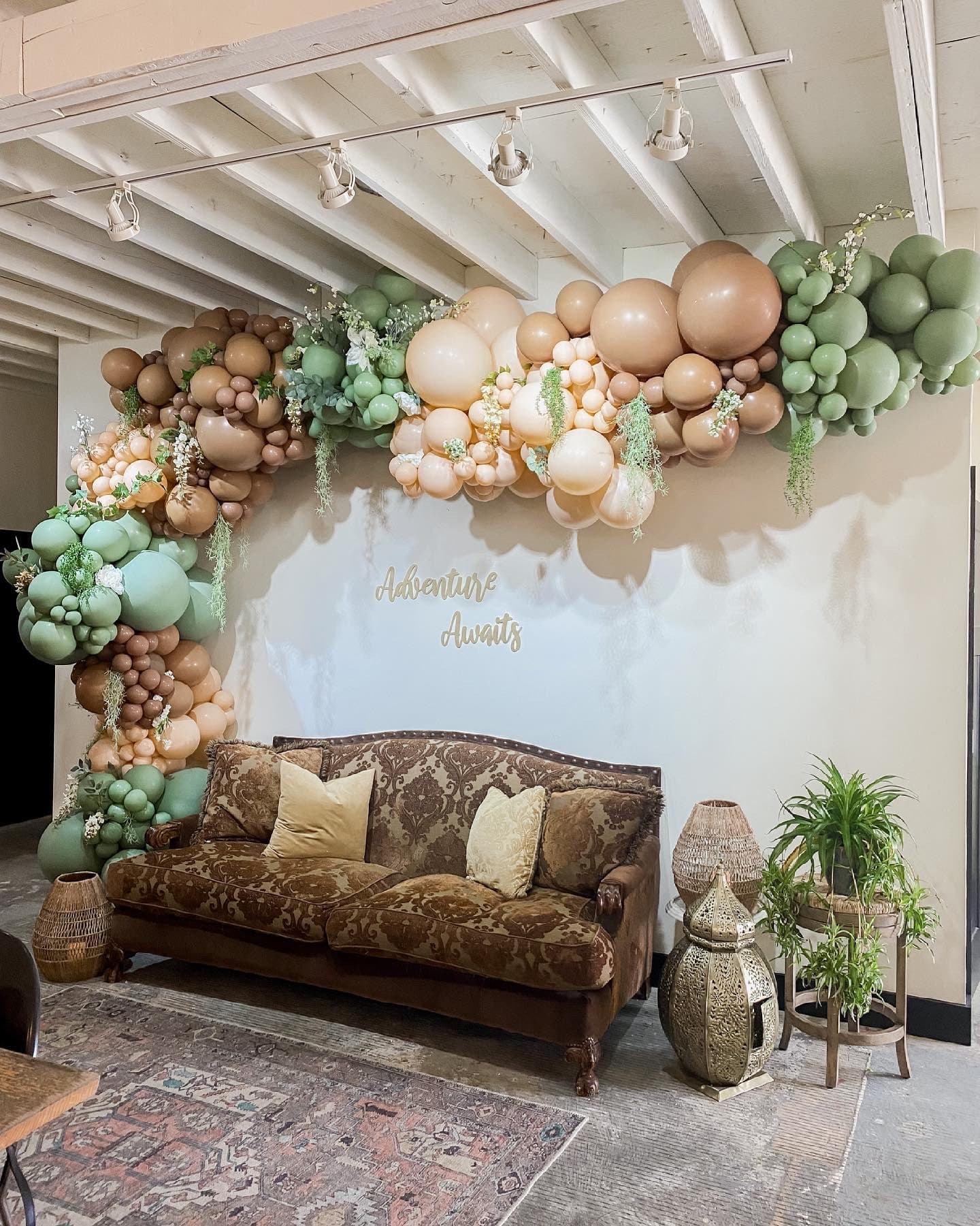 Adventure Awaits, an organic balloon garland in sage, sand, and clay arcs over a vintage brown damask settee with gold pillows, a Moroccan brass lantern, and a potted palm, beneath a gold 'Adventure Awaits' script.