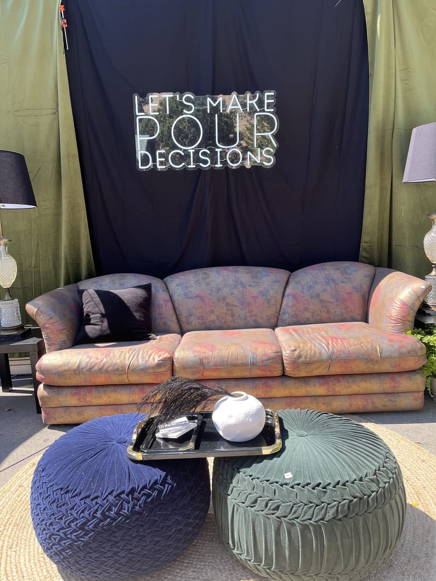 A festival VIP lounge, a rust-and-blush velvet sofa with a navy and a sage bouclé pouf on a layered rug, against a black backdrop reading 'Let's Make Pour Decisions'.