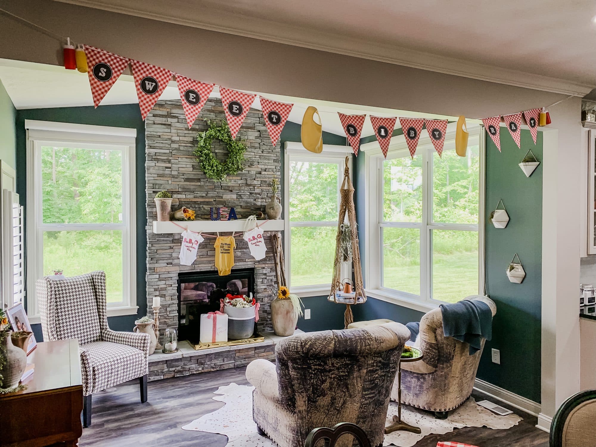 A styled sunroom for a BBQ baby shower, red gingham 'Sweet Baby' bunting strung across windows above a stone fireplace, with a cowhide rug and plaid wingback chairs.