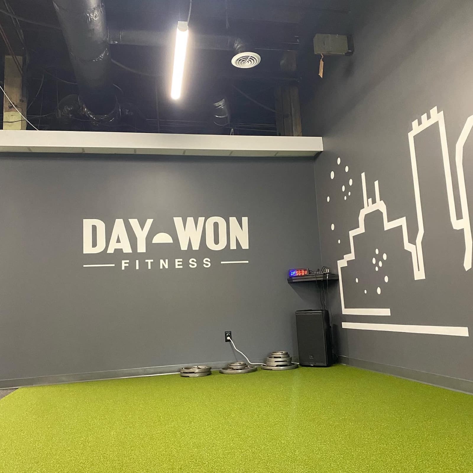 A hand-painted white Day Won Fitness wordmark and a minimal city skyline running across a charcoal gym wall above bright green turf.
