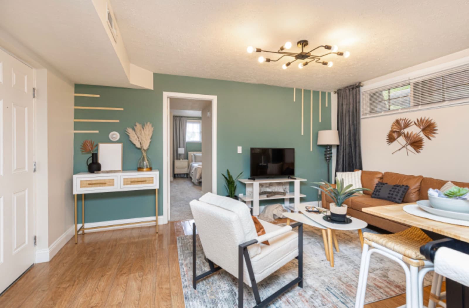 Delaware St. living room after staging, sage accent wall, tan leather sofa, bouclé chair, and a brass linear chandelier.