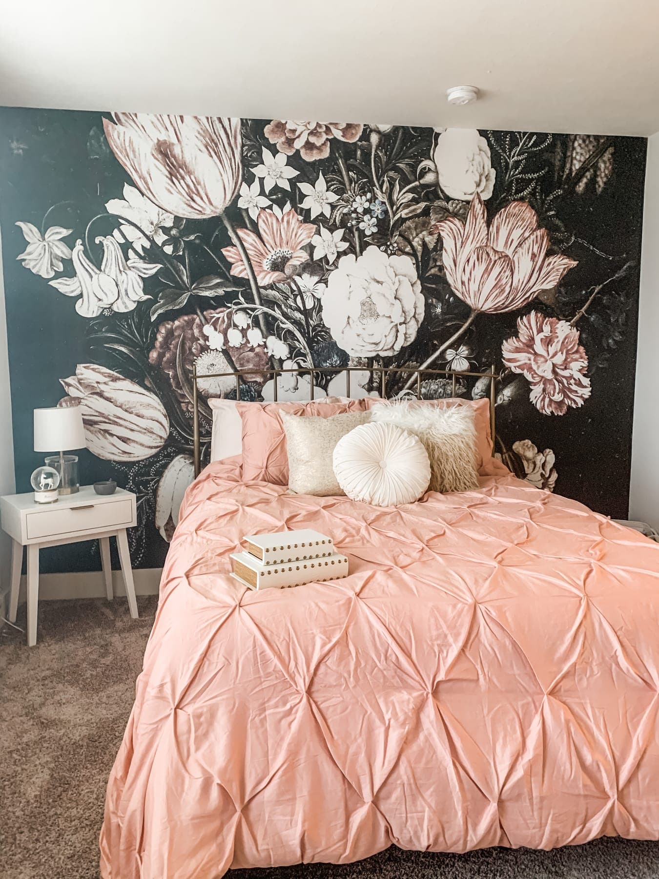 A teen bedroom with a dark floral mural wall behind the bed, blush pintuck bedding with white faux-fur pillows, and a white nightstand with a small lamp.
