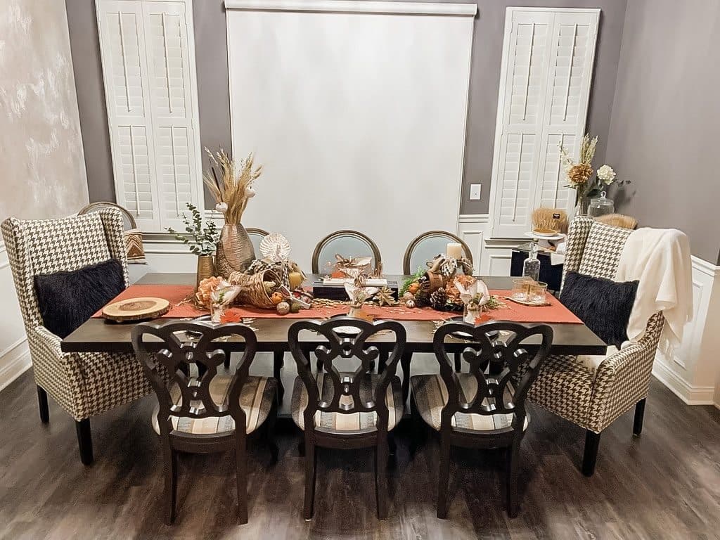 A Friendsgiving table set in burnt orange and copper, a cornucopia spilling wheat and dried florals down the center, between houndstooth wingbacks and dark carved chairs.