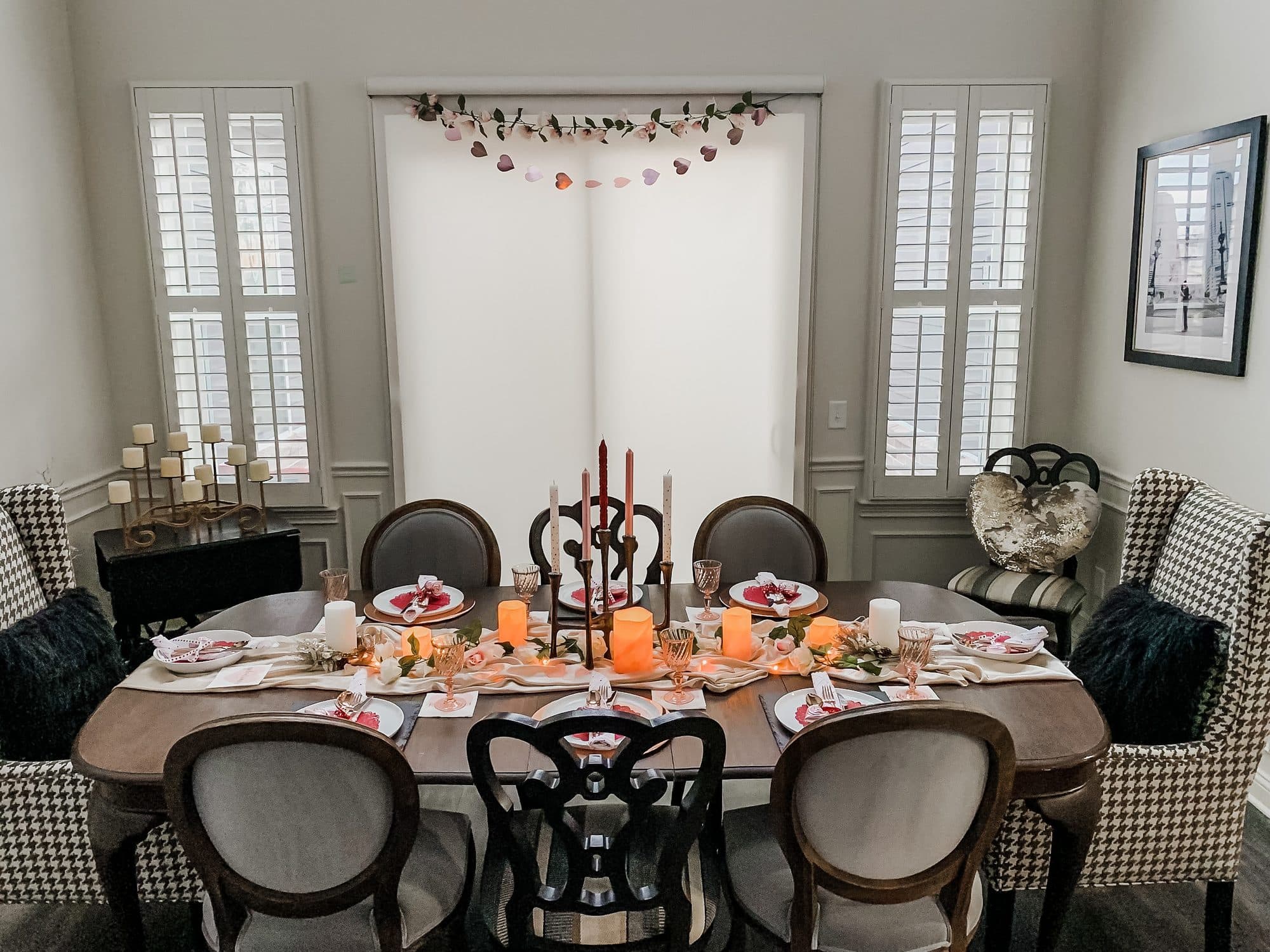 A Galentine's table under a crystal chandelier, heart place settings, taper and pillar candles, and a gauzy runner, framed by houndstooth wingbacks.