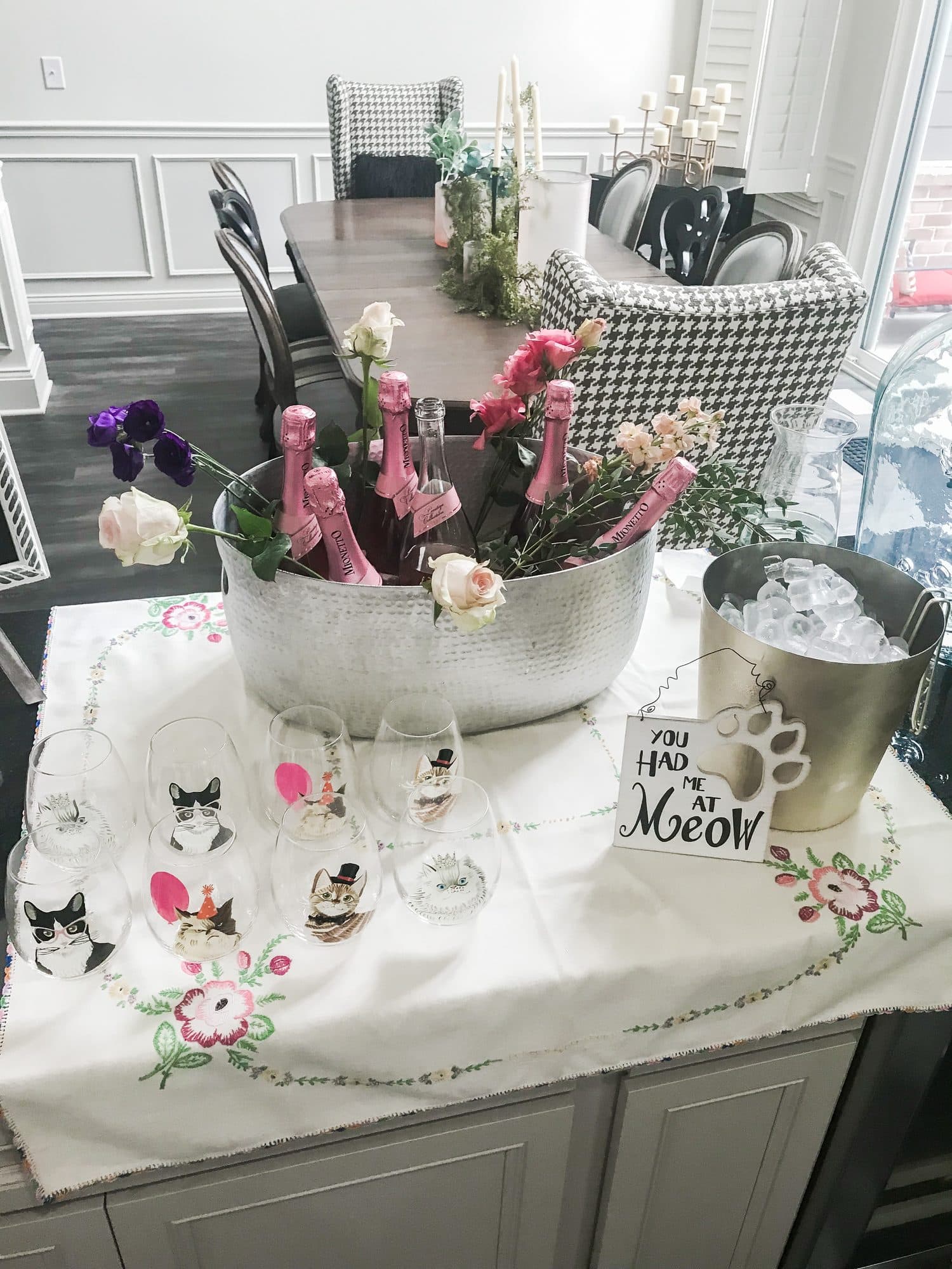 A bridal-shower drink station, a hammered ice bucket of rosé and roses with cat-face wine glasses on an embroidered cloth, beside a 'You Had Me at Meow' sign.