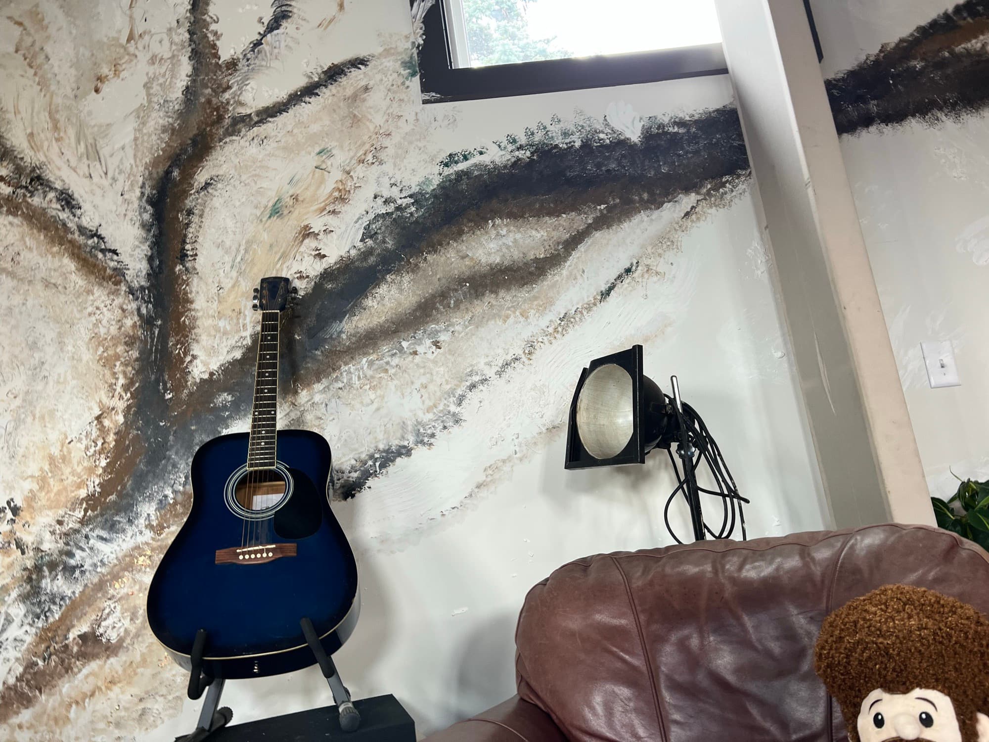 A floor-to-ceiling hand-painted abstract feature wall in bone, ink, and metallic browns, with a blue acoustic guitar on a stand in front of it.
