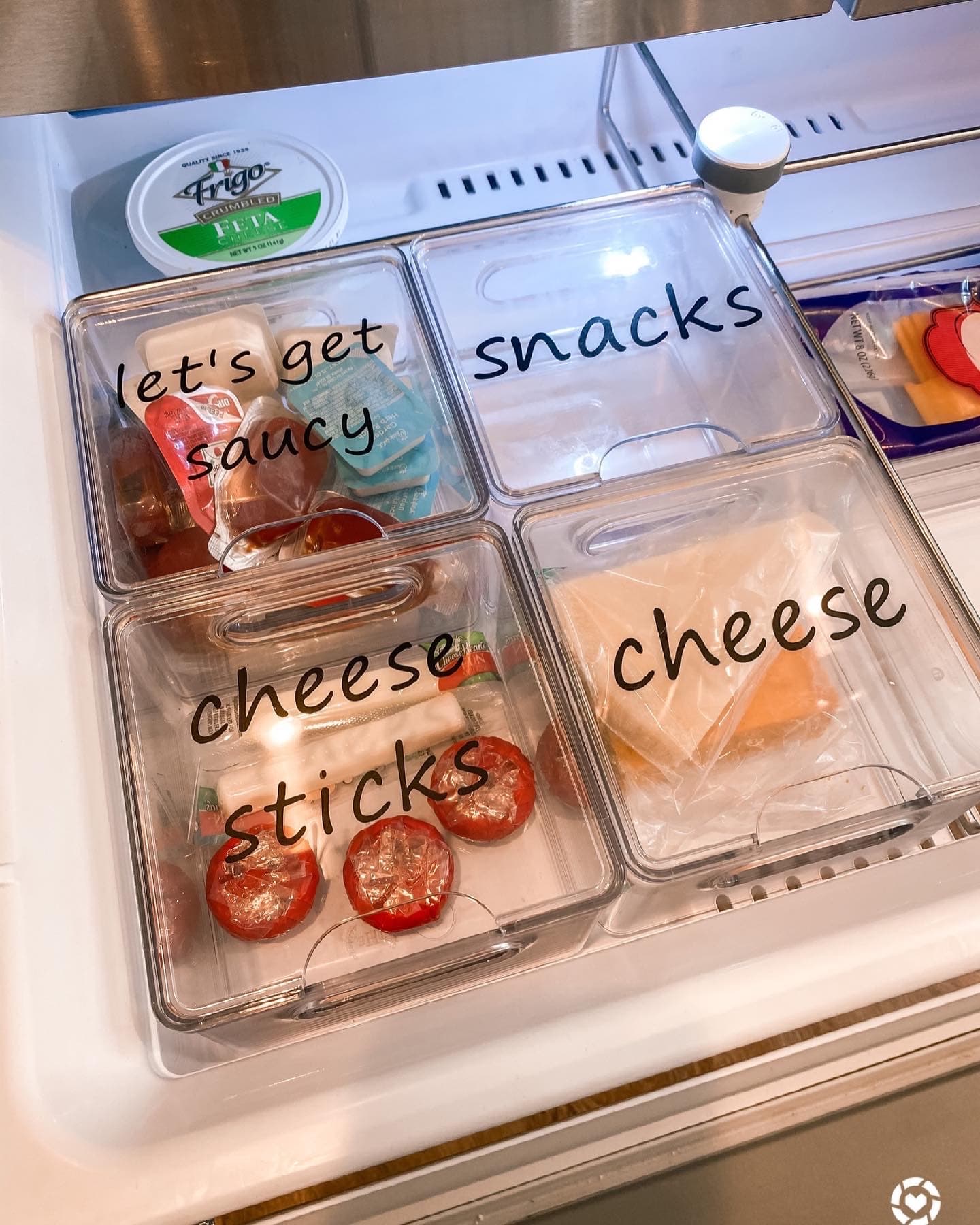 Labeled clear bins reading 'snacks', 'cheese', and 'let's get saucy' in an organized fridge drawer.