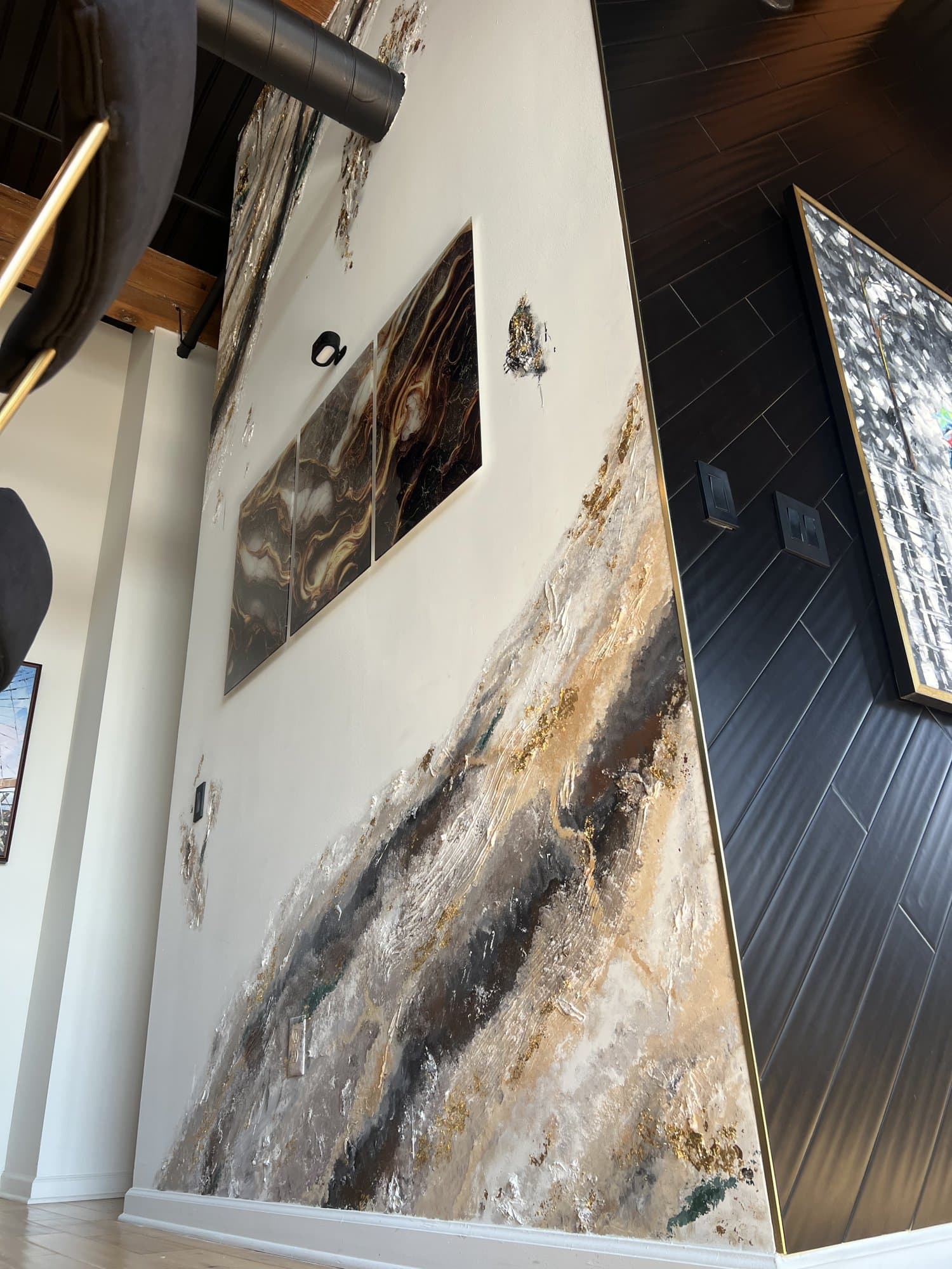 Levels after, a floor-to-ceiling feature wall of poured gold, black, and cream with a brass reveal, framed art inset, beside dark slat paneling.