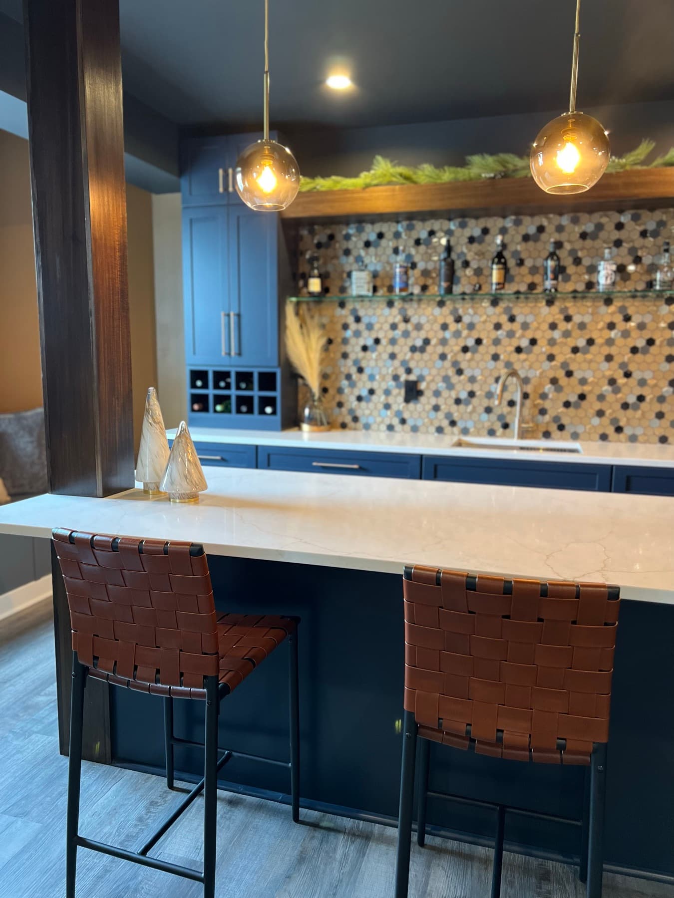 After, a dark hosting lounge with a backlit decorative screen wall, a walnut bar with a brass rail, and woven leather stools.