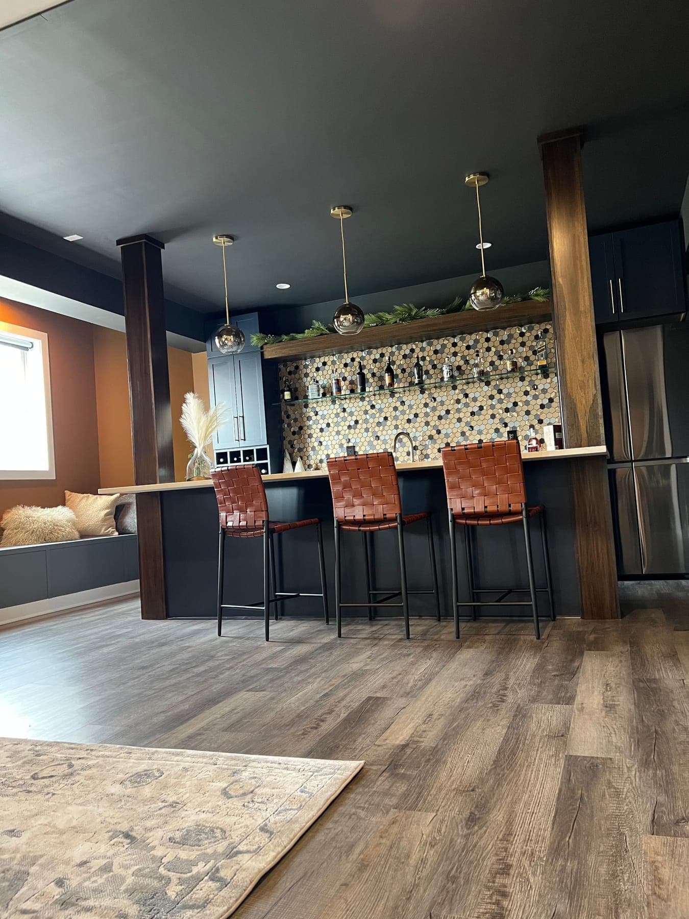 After, the finished lounge from the seating side: dark walls, a backlit hex-tile bar wall under brass pendants, woven leather stools, and a patterned rug on wide-plank floors.