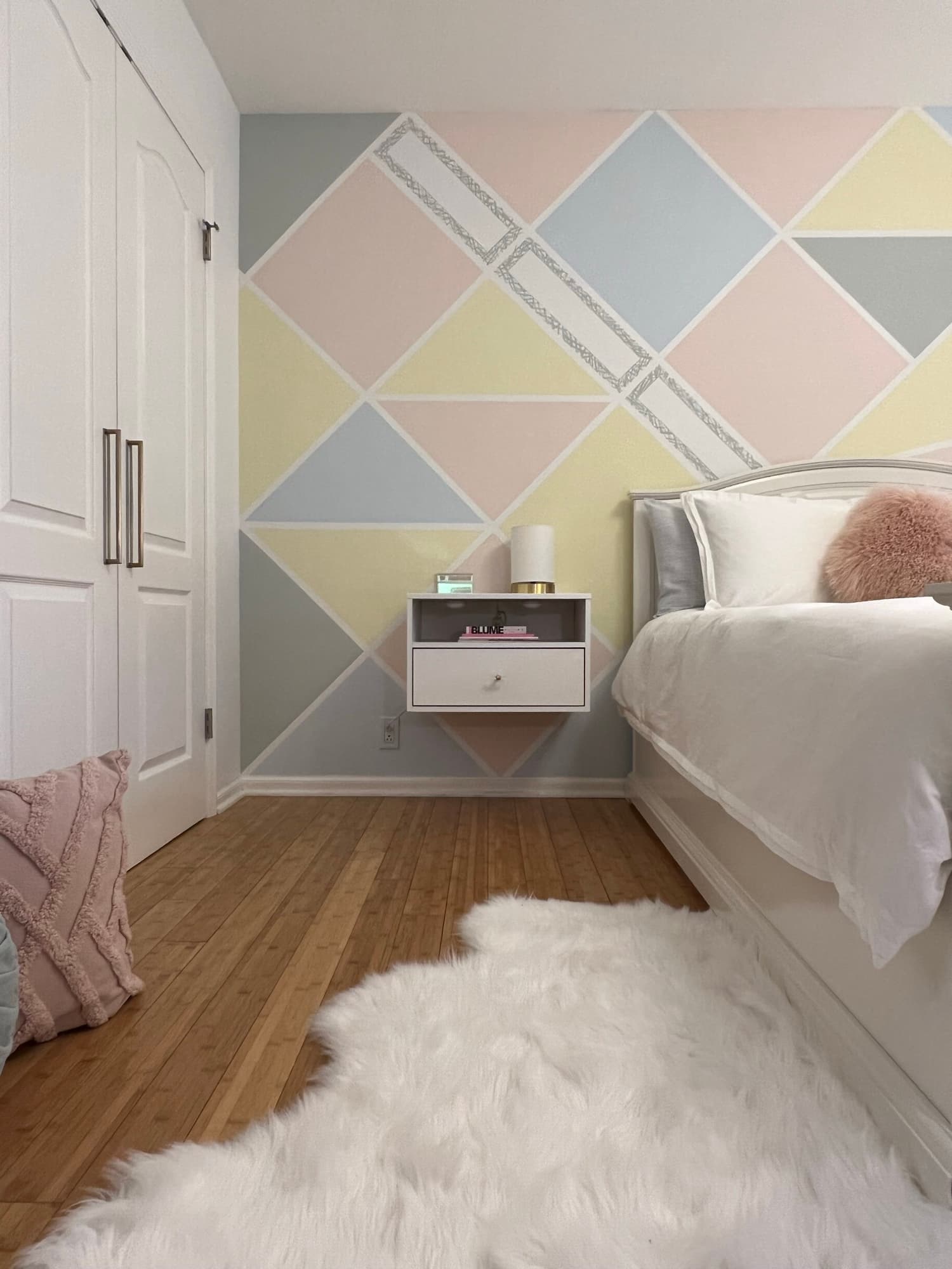 A teen bedroom with a hand-painted diamond accent wall in blush, butter yellow, and dove gray, a white bed with a blush faux-fur pillow, a floating nightstand, and a sheepskin rug over bamboo flooring.