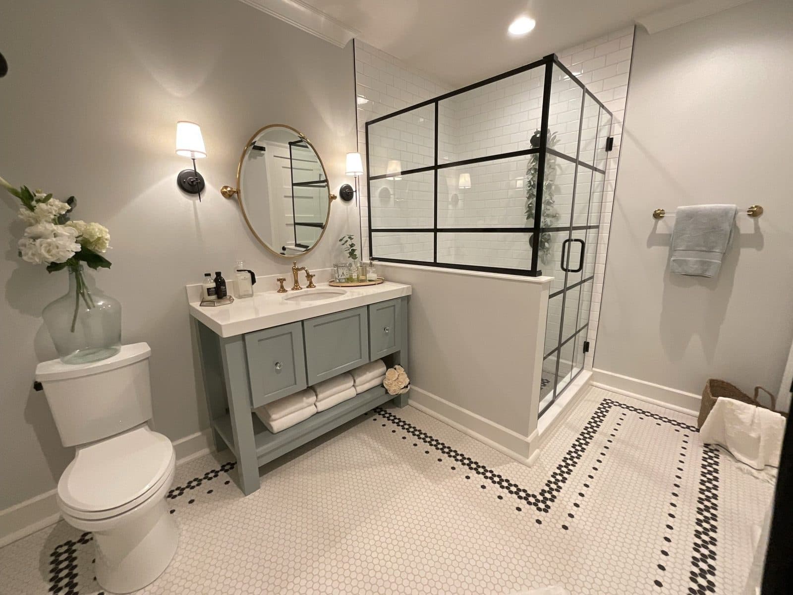 A full bath with a sage-blue vanity, a round brass-rimmed mirror, a black-framed glass shower, and white hex tile with a black Greek-key border.