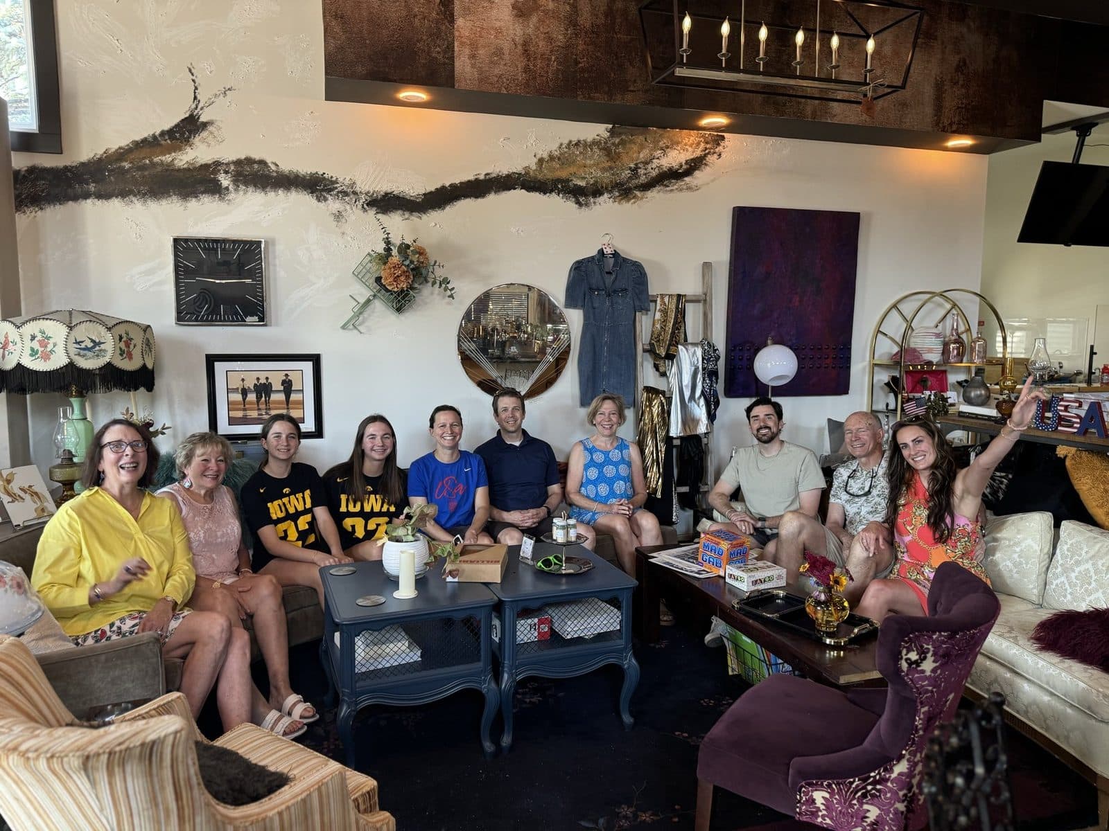 The Skōsh living room full of people: a dozen regulars on the sofas and vintage chairs under the branch installation, sunlight through the storefront glass.