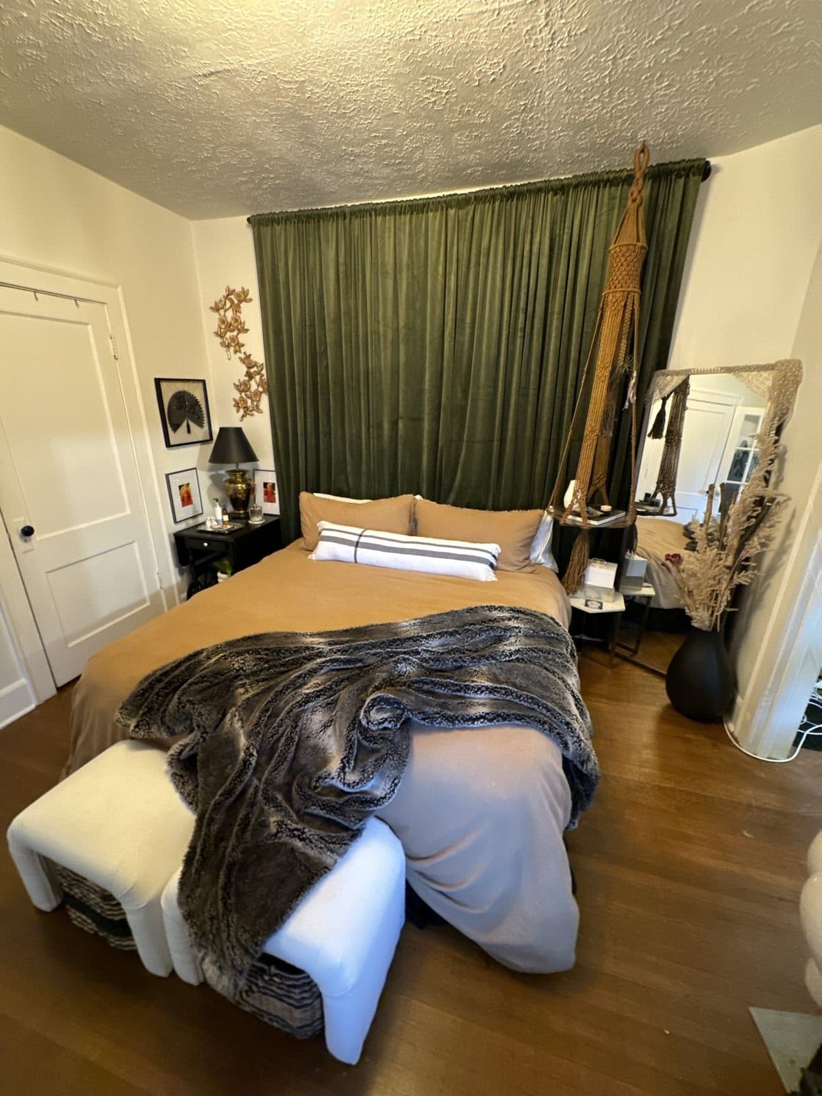 A small bedroom made to feel like a hotel: olive velvet drapes floor to ceiling behind the bed, camel linen, a faux-fur throw, a white bench, and a black cat on the pillows.