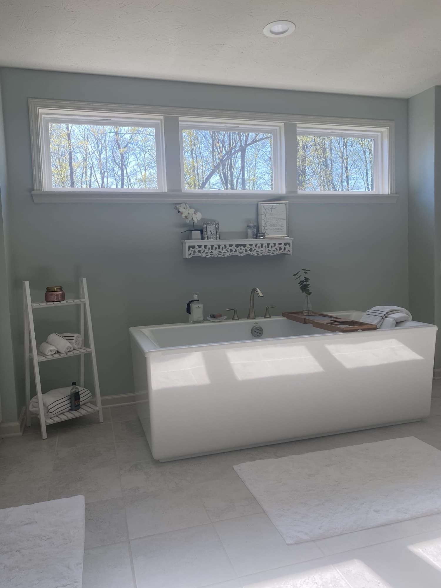 A freestanding soaking tub against sage-green walls, beneath a bank of three windows onto the trees, with a wood bath caddy, towels, and an orchid on a wall shelf.