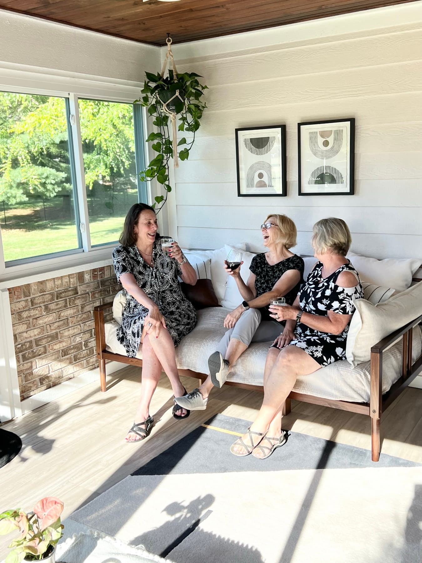 Three friends laughing over wine on the linen daybed beneath the gallery wall, pothos trailing from the window and the yard bright behind them.